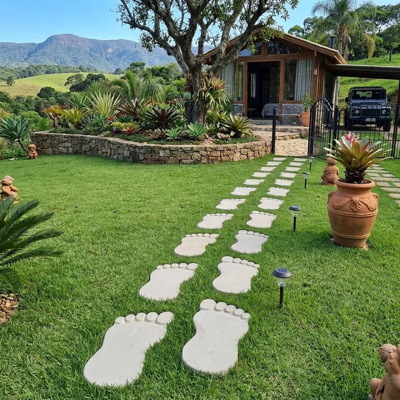 Caminho de pisantes em formato de pés no gramado, com luminárias baixas e uma casa ao fundo.