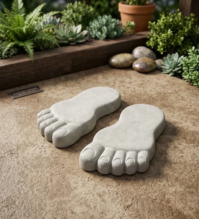 Duas peças de concreto em formato de pés sobre o solo, com plantas e pedras ao fundo.