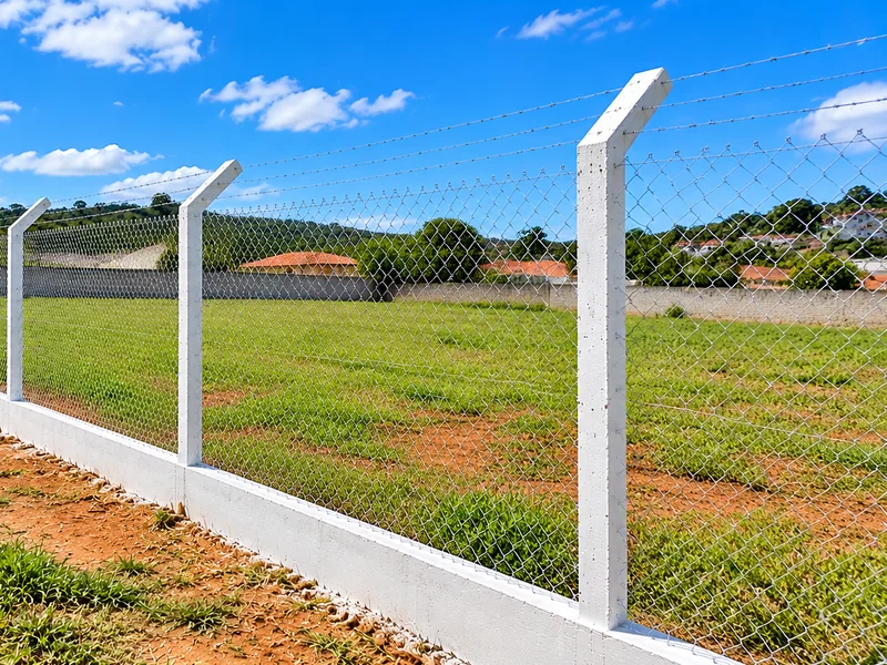 Mourões de concreto sustentando tela de alambrado em um terreno com casas ao fundo